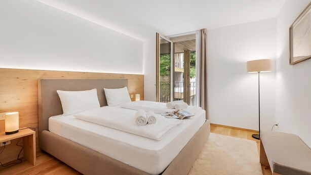 Ein ordentlich bezogenes Bett mit weißen Leinen und Kissen, eine Nachttischlampe und Blick ins Freie durch ein Fenster.