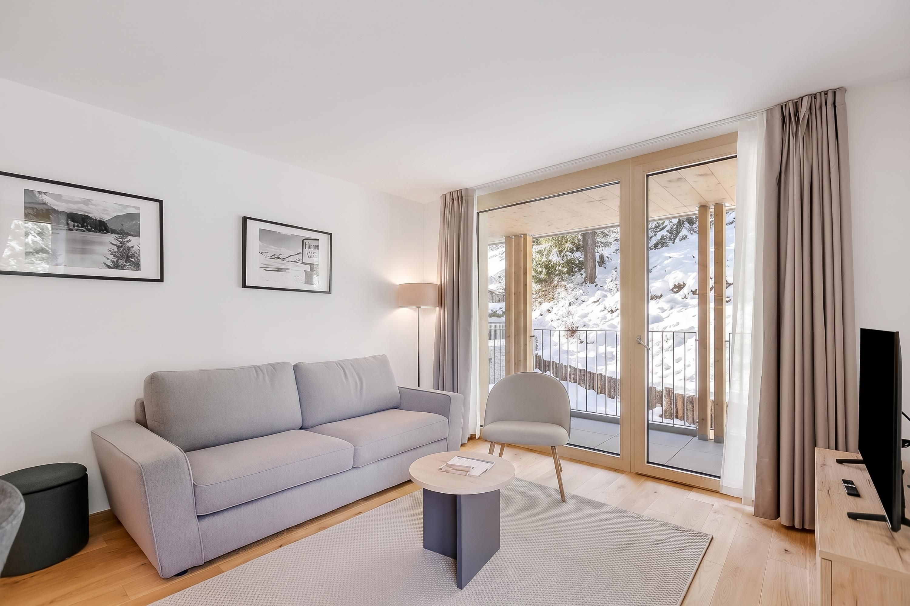 Un saló modern amb un sofà gris, una taula de centre rodona i un gran finestral corredís que dona a un balcó amb vistes a la neu.