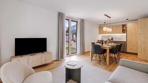 Ein modernes Wohnzimmer mit Flachbildfernseher, eine Essecke mit Tisch für zwei Personen und eine Küche mit Holzschränken und Pendelleuchten.