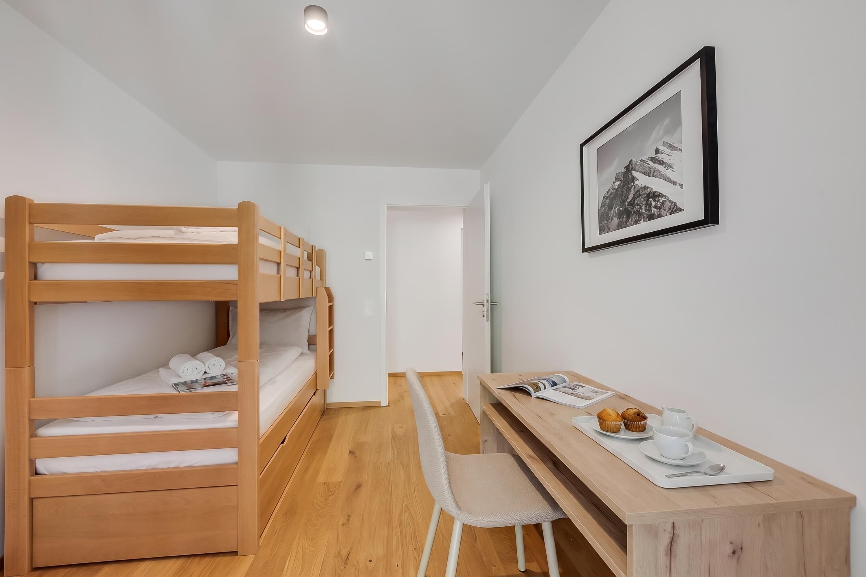 Una habitació amb un llit superposat, un escriptori amb una cadira, un quadre al mur i una porta que condueix a una altra habitació.