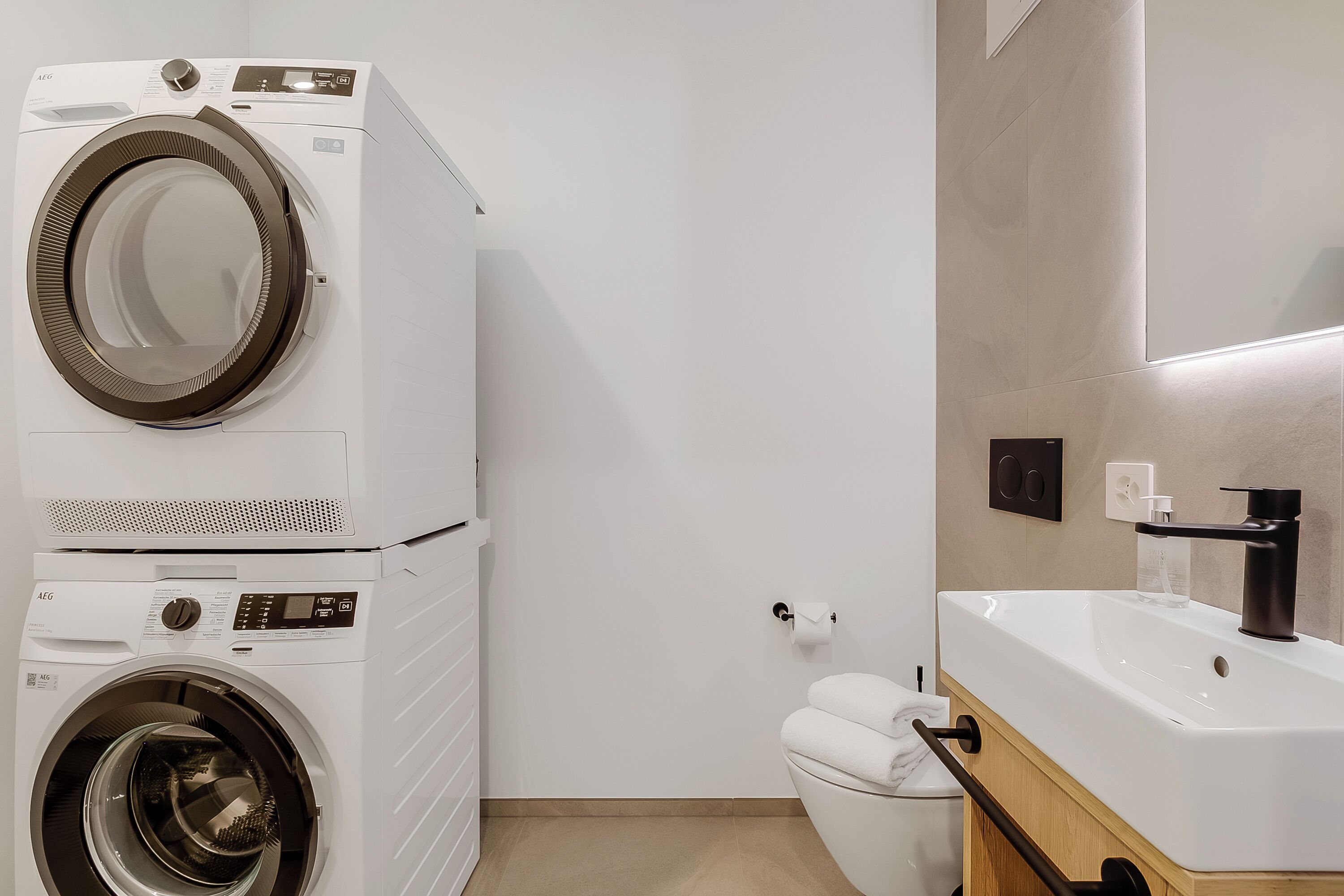 Ein kleiner Waschraum mit einer Waschmaschine und einem Trockner mit Frontlader, einer Toilette und einem Waschbecken mit modernem Wasserhahn.