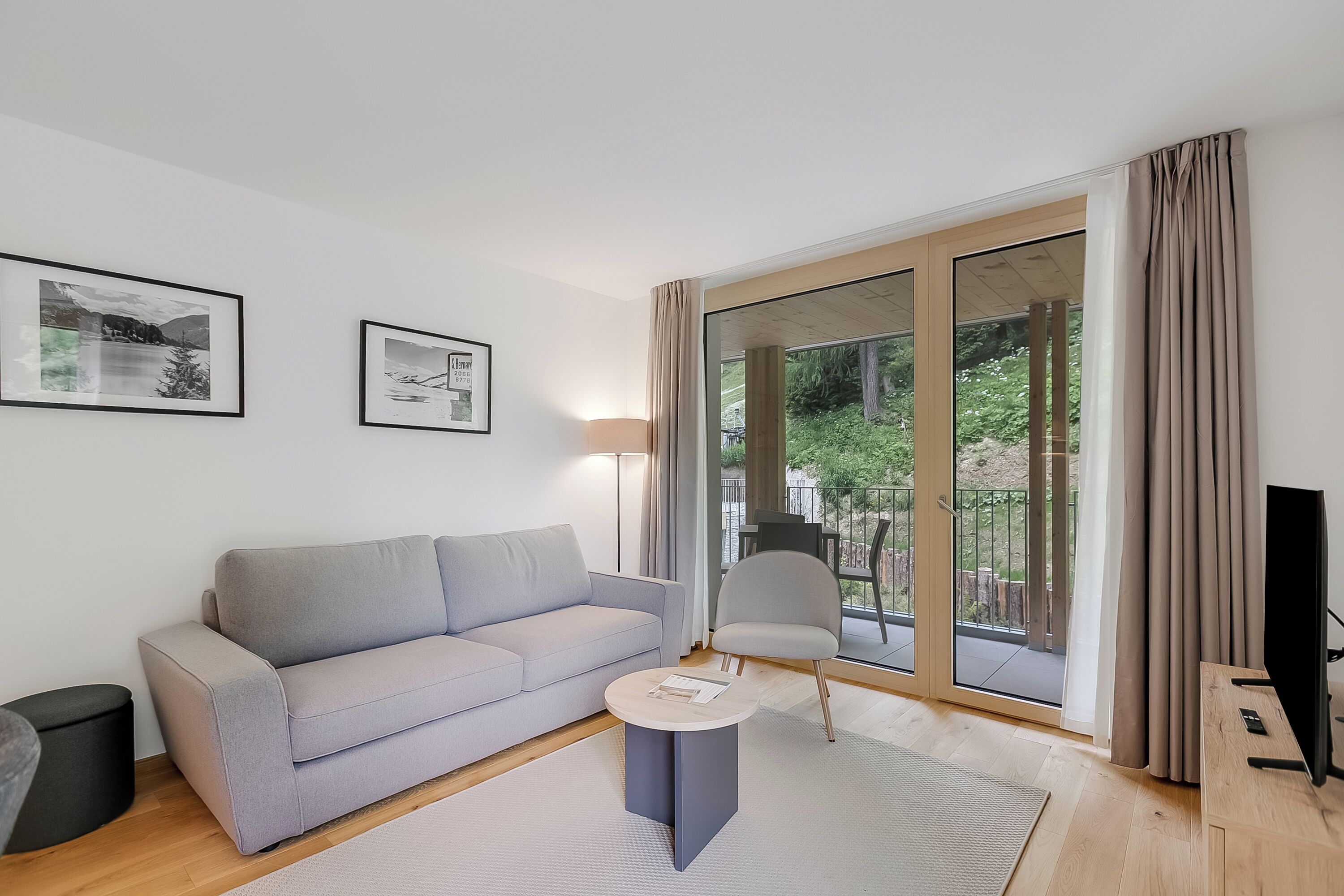 Ein modernes Wohnzimmer mit einem grauen Sofa, einem kleinen runden Tisch und einem Fernsehschrank. An der Wand hängen zwei gerahmte Bilder, und ein großes Schiebeglastür führt zu einem Außenbereich.