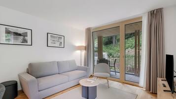 Ein modernes Wohnzimmer mit einem grauen Sofa, einem kleinen runden Tisch und einem Fernsehschrank. An der Wand hängen zwei gerahmte Bilder, und ein großes Schiebeglastür führt zu einem Außenbereich.