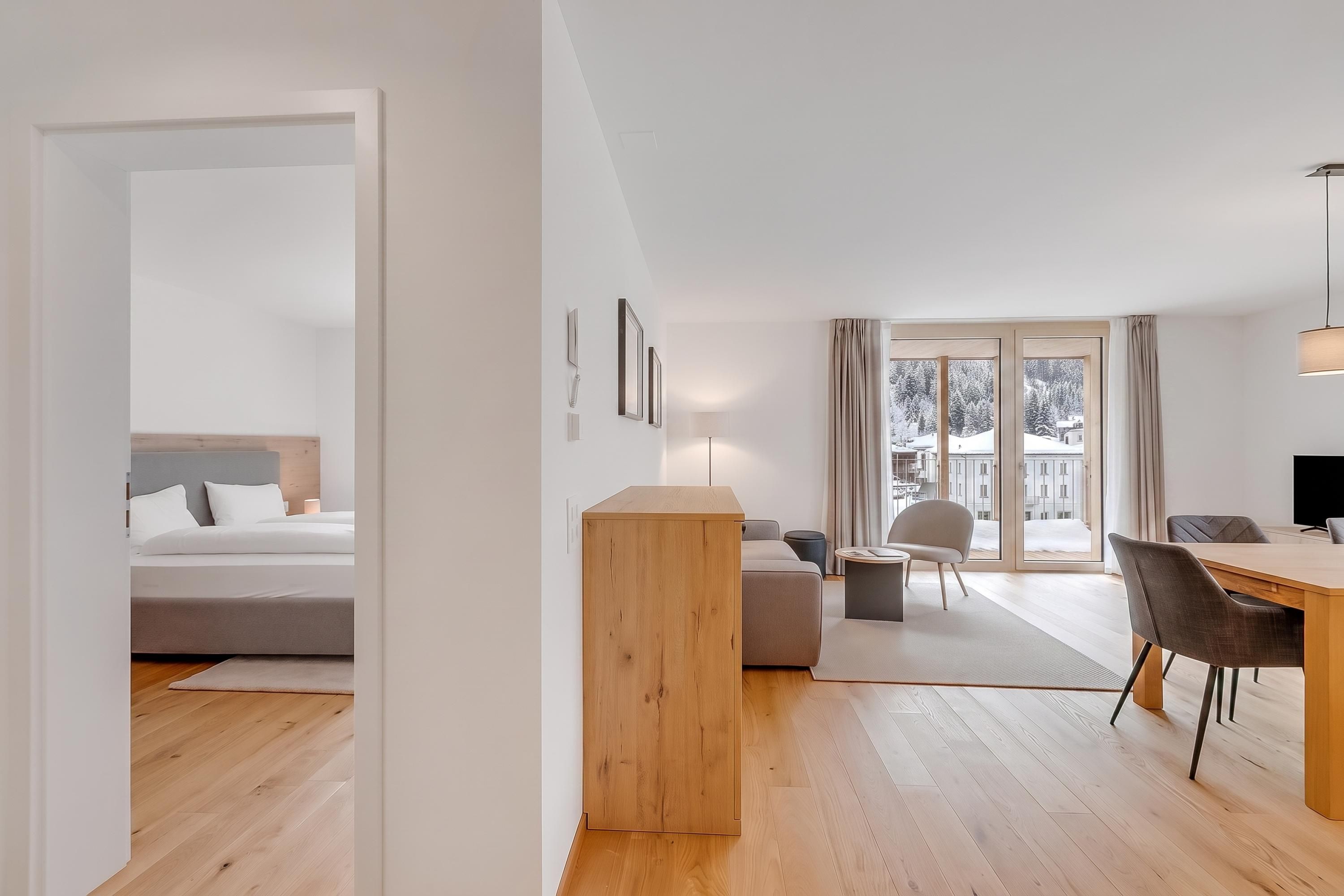 Una habitació d'hotel moderna amb un llit, una taula auxiliar de fusta, un sofà, una taula de menjador amb cadires i una finestra gran amb vista a un paisatge nevàt.