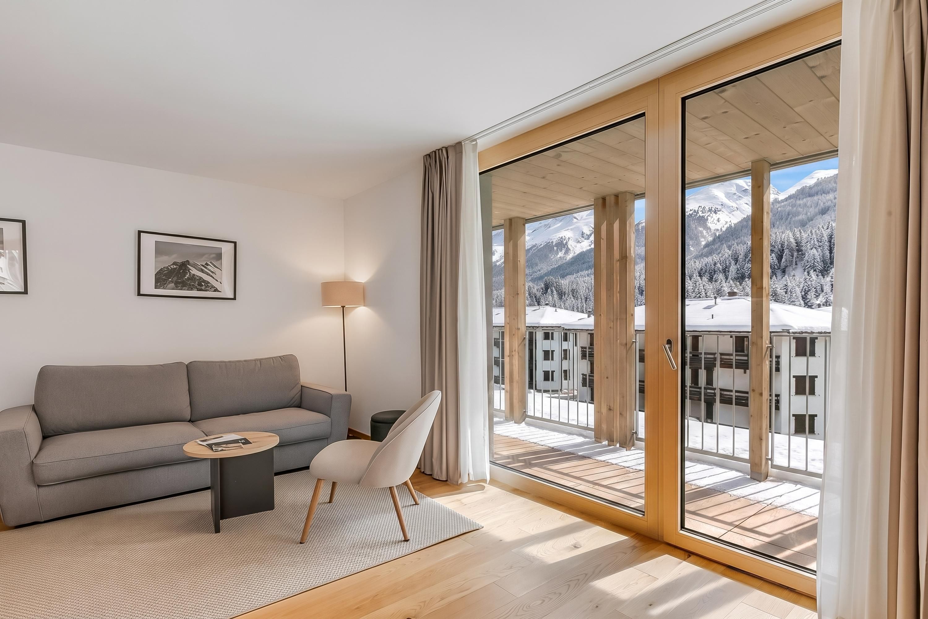 A modern living room with a grey sofa, a small round table, and a chair. There are two framed pictures on the wall, a floor lamp, and a large sliding glass door leading to a balcony with a snowy mountain view.