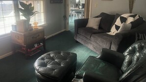 Ein Wohnzimmer mit einem dunkelgrauen Sofa, einem Ledersitzhocker und einem Couchtisch. An der Wand hängt ein gerahmtes Bild und auf dem Fensterbrett steht eine Pflanze.