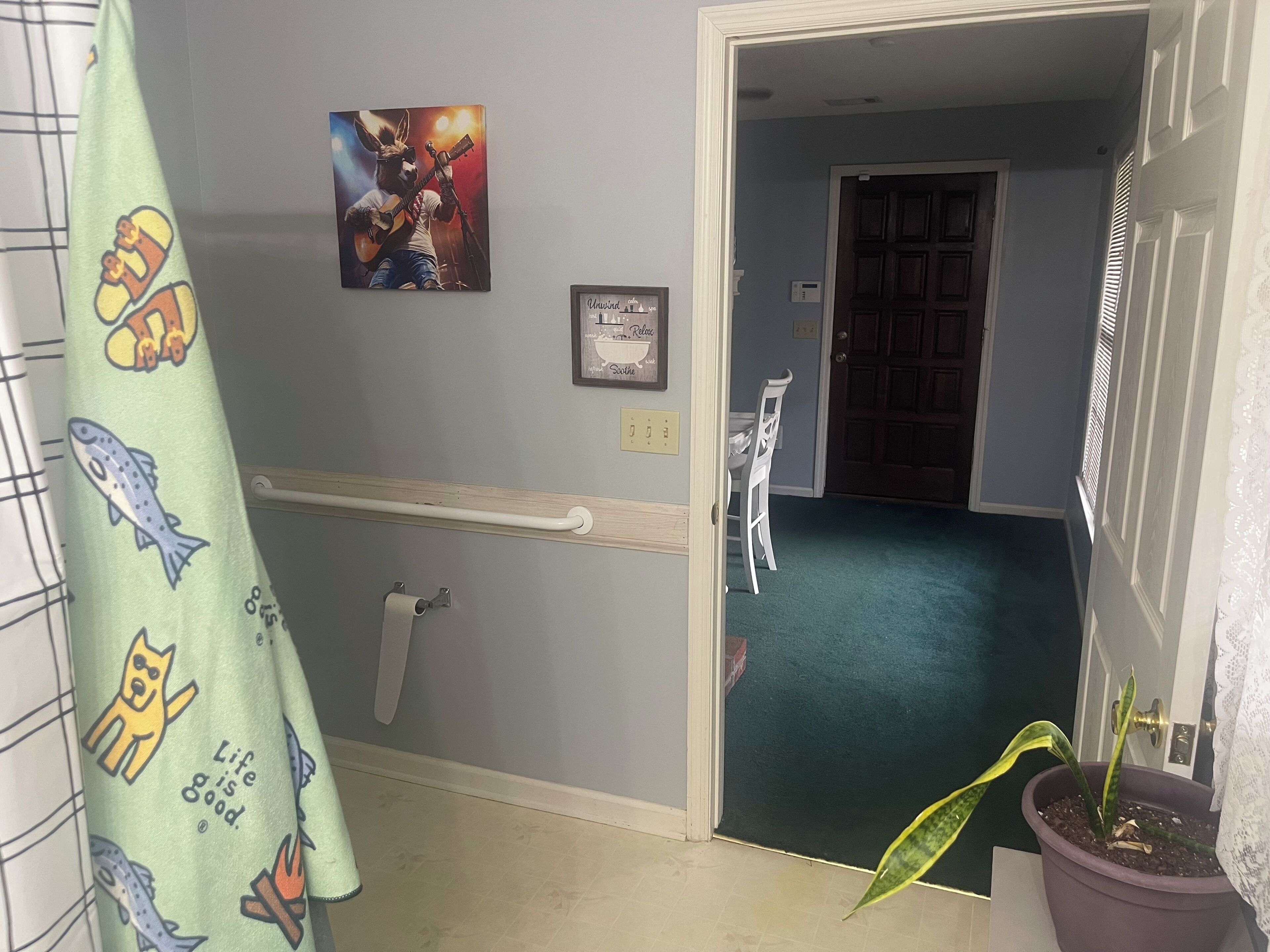 A hallway with a handrail, a door leading to a room, a potted plant, and a wall with a framed picture and a poster.
