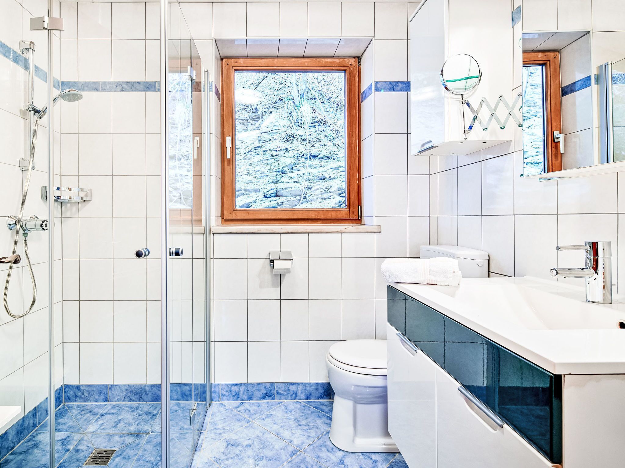 Een moderne badkamer met een glazen douchecabine, een toilet, een wastafel met spiegel en een raam met uitzicht op een besneeerd landschap.