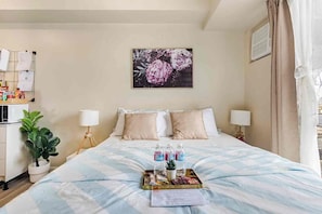 A hotel room with a bed, two bedside lamps, a wall-mounted air conditioner, a bulletin board, and a painting of flowers. - Studio Lune | Modern Makati Studio | Queen Bed | Fast Wi-Fi | Self Check-in ⭐ (Makati City)