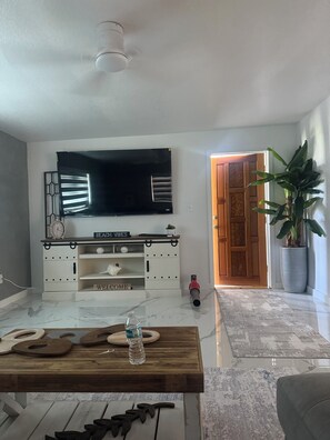 A living room with a flat-screen TV mounted on a white cabinet, a wooden coffee table with a water bottle, and a potted plant. - Charming 3-bedroom house with pool in beautiful Palm Bay (Palm Bay)