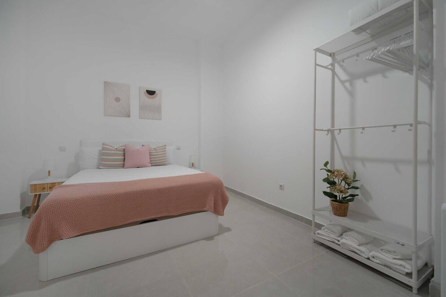 Ein schlicht eingerichtetes Schlafzimmer mit Bett, Nachttisch, Wandregal und einer Pflanze.