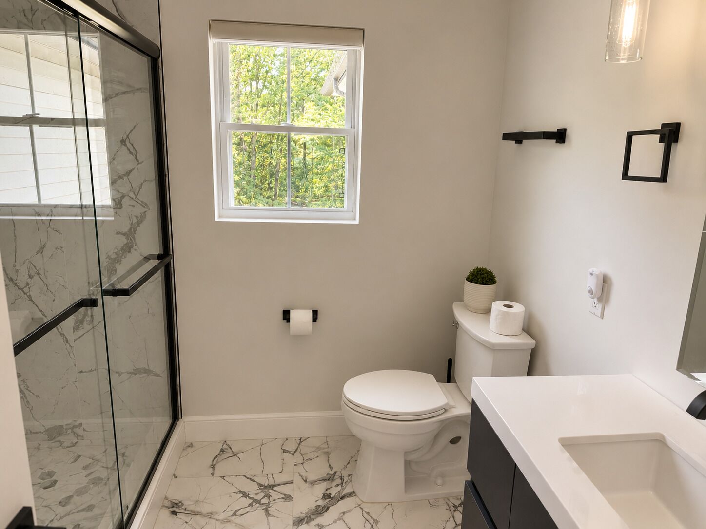 A bathroom with a glass shower enclosure, a toilet, a sink, and a window with a view of greenery.