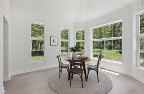 Uma sala de jantar com uma mesa redonda, quatro cadeiras e vista para uma floresta através de grandes janelas.