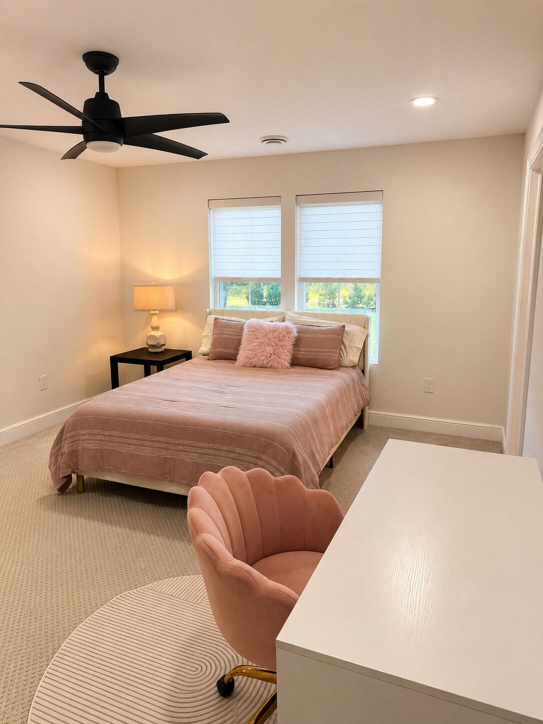 A bedroom with a bed, a desk, a chair, a ceiling fan, and a window with blinds.