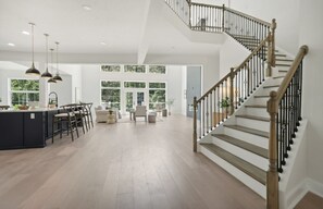 Uma sala de estar espaçosa com uma ilha central, uma escada com corrimão de madeira e grandes janelas que proporcionam luz natural.