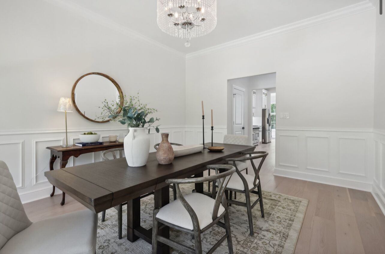 A dining room with a wooden table, white upholstered chairs, a chandelier, a round mirror, and a vase with flowers.