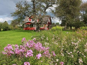 A red house with a sloped roof surrounded by a lush green field and flowering plants. - Schönes Ferienhaus in Småland mit Sauna, Bootssteg zum Schwimmen mit Ruderboot by Interhome (Tingsryd)