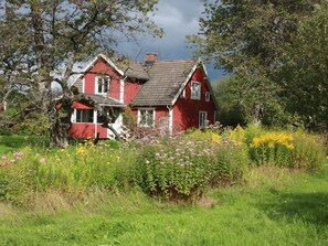 A red house with a sloped roof, surrounded by a garden with various plants and flowers. - Schönes Ferienhaus in Småland mit Sauna, Bootssteg zum Schwimmen mit Ruderboot by Interhome (Tingsryd)