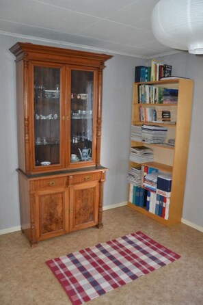 A room with a wooden cabinet with glass doors, a bookshelf filled with books and binders, and a plaid rug on the floor. - Schönes Ferienhaus in Småland mit Sauna, Bootssteg zum Schwimmen mit Ruderboot by Interhome (Tingsryd)