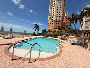 A swimming pool with a curved design, surrounded by a brick deck and palm trees. - Cape Marco Paradise: Beachfront Monterrey Condo, #302 with 2 Balconies (Marco Island)