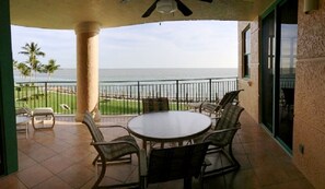 A covered outdoor area with a round table and chairs, a view of the ocean, and a palm tree in the background. - Cape Marco Paradise: Beachfront Monterrey Condo, #302 with 2 Balconies (Marco Island)