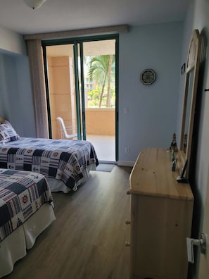 A bedroom with two beds, a wooden dresser, a mirror, and a sliding glass door leading to a balcony with a view of palm trees. - Cape Marco Paradise: Beachfront Monterrey Condo, #302 with 2 Balconies (Marco Island)