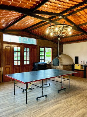 Una mesa de ping pong en una habitación con techo rústico, puertas de madera y un horno de piedra.