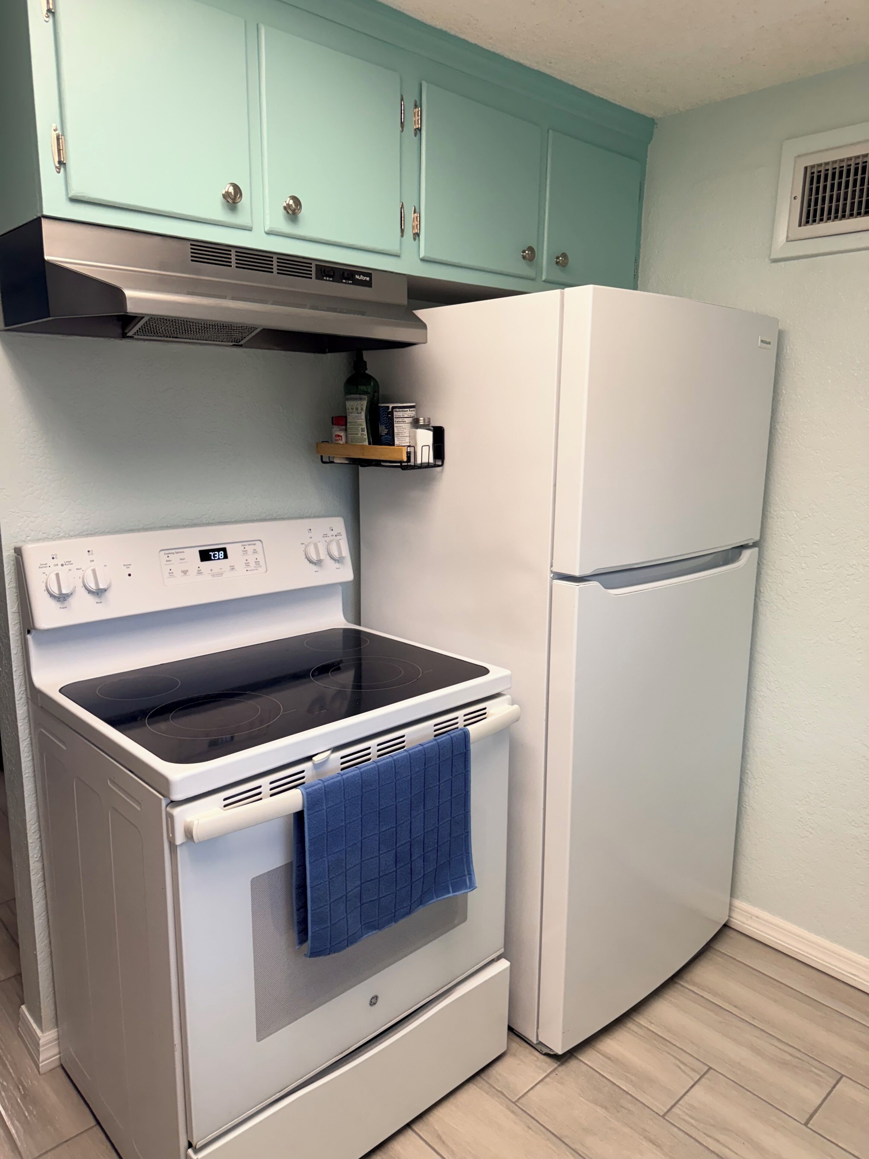 A kitchen with a white stove, a white refrigerator, teal cabinets, and a small shelf with kitchen items.