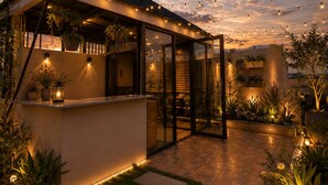 Un área de estar al aire libre techada, con luces colgantes, plantas en macetas y un sendero que lleva a la entrada de un edificio.