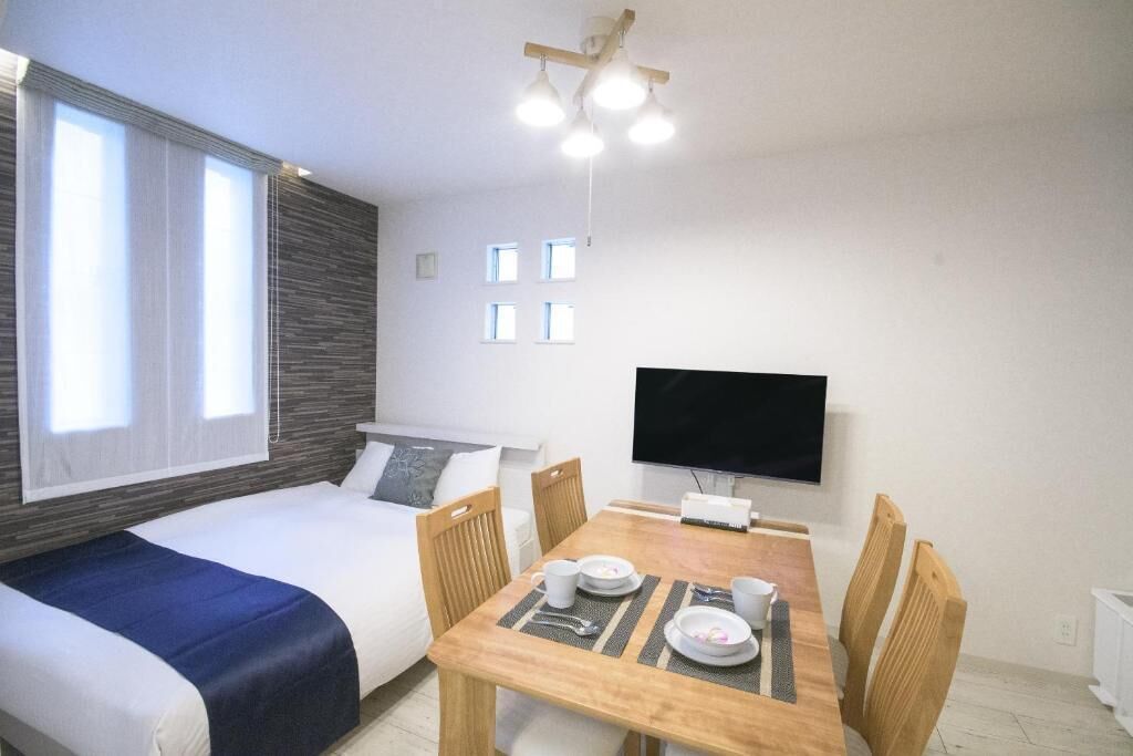 A room with a bed, a dining table with chairs, a flat-screen TV, and a window with curtains.