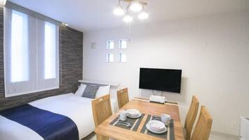A room with a bed, a dining table with chairs, a flat-screen TV, and a window with curtains.