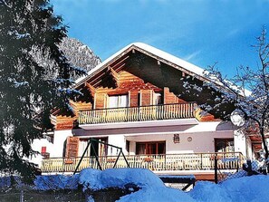 En tvåvånings chalet med en balkong, omgiven av snötäckta träd och berg.