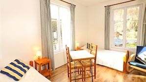 A room with a bed, a table with chairs, a lamp, and a view of the outdoors through the windows.