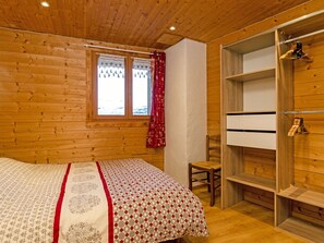 Quarto de cabana de madeira com cama, uma cadeira, um guarda-roupa e uma janela com cortinas.