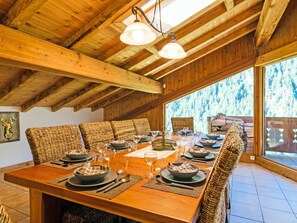 Ein Essbereich mit Holzboden und hohen Decken, ein langer Tisch, der für eine Mahlzeit gedeckt ist, und Blick auf einen Wald durch große Fenster.