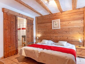 A bedroom with a wooden ceiling, a bed with white bedding and a red blanket, a nightstand with a lamp, and a framed botanical illustration on the wall. - Chalet Honore by Interhome (Peisey-Nancroix)
