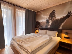 Ein Hotelzimmer mit einem großen Bett, einem Kopfteil mit einer Berglanschaftsmalerei, einem Nachttisch mit einer Lampe und durchsichtigen Vorhängen am Fenster.