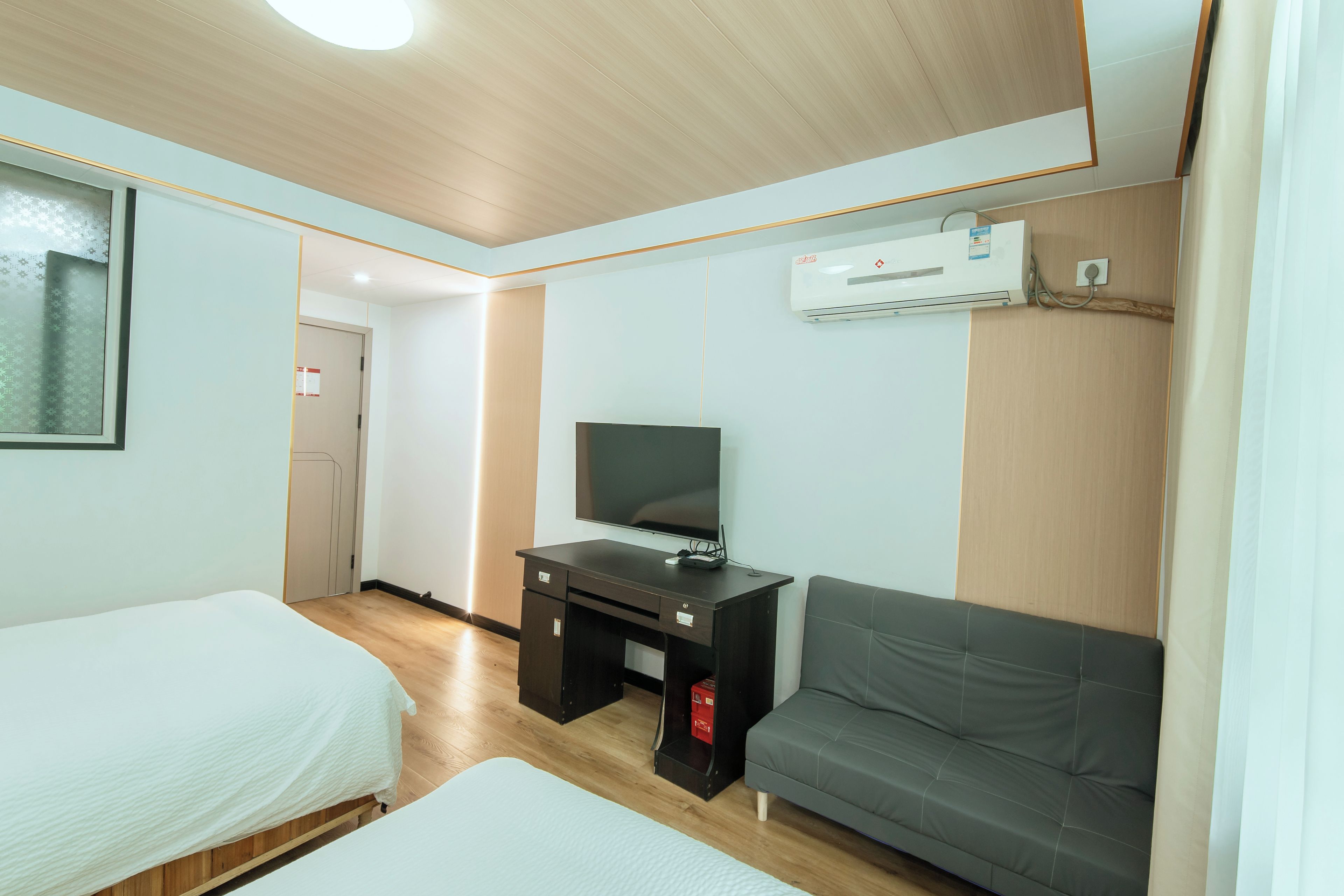 A hotel room with two beds, a desk with a television, a grey sofa, and an air conditioning unit.