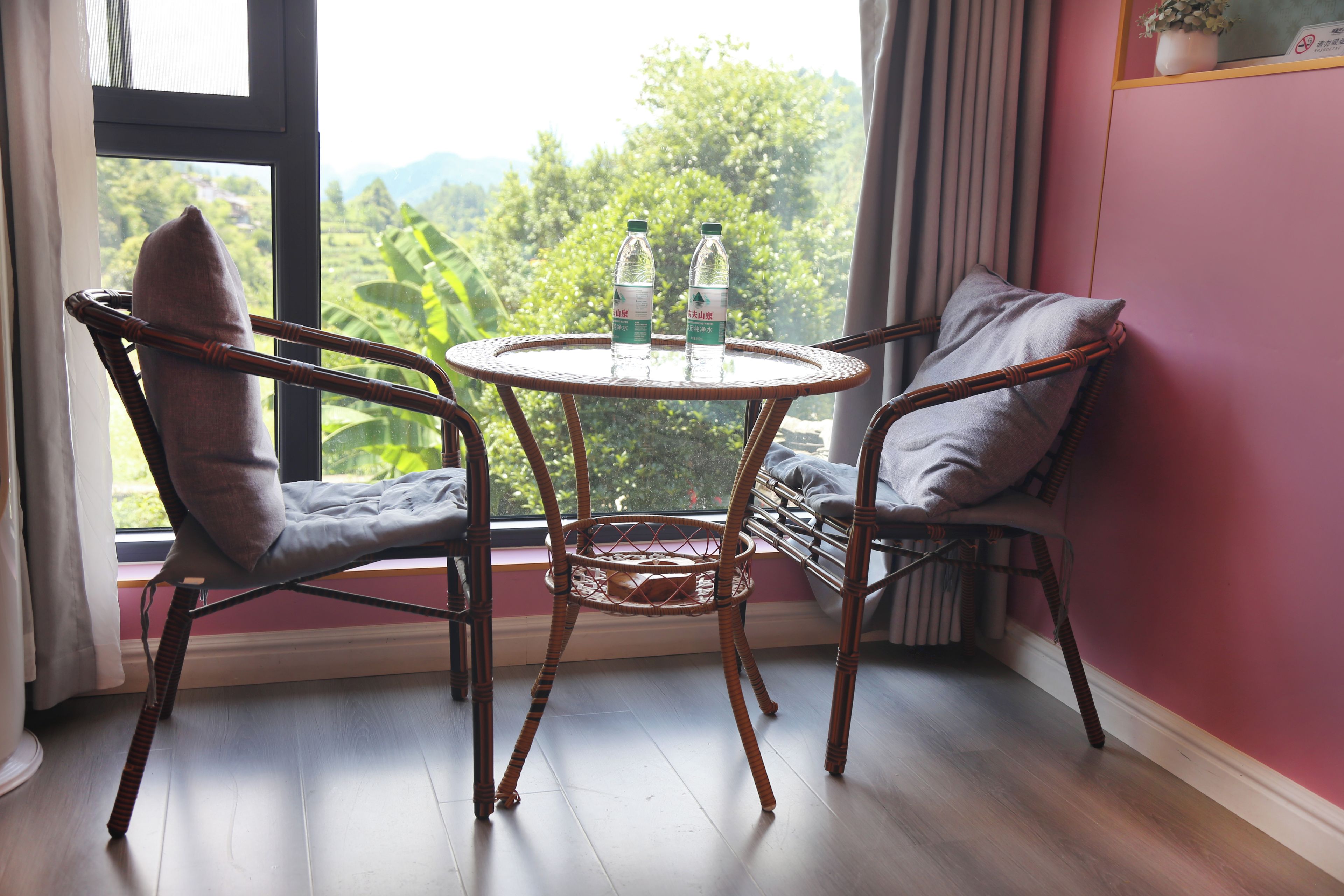 Quarto com uma mesa redonda, duas cadeiras de vime, uma janela com vista para vegetação e um vaso de planta no parapeito.