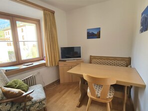A room with a wooden table, a television on a stand, a chair, a radiator, and a window with curtains.