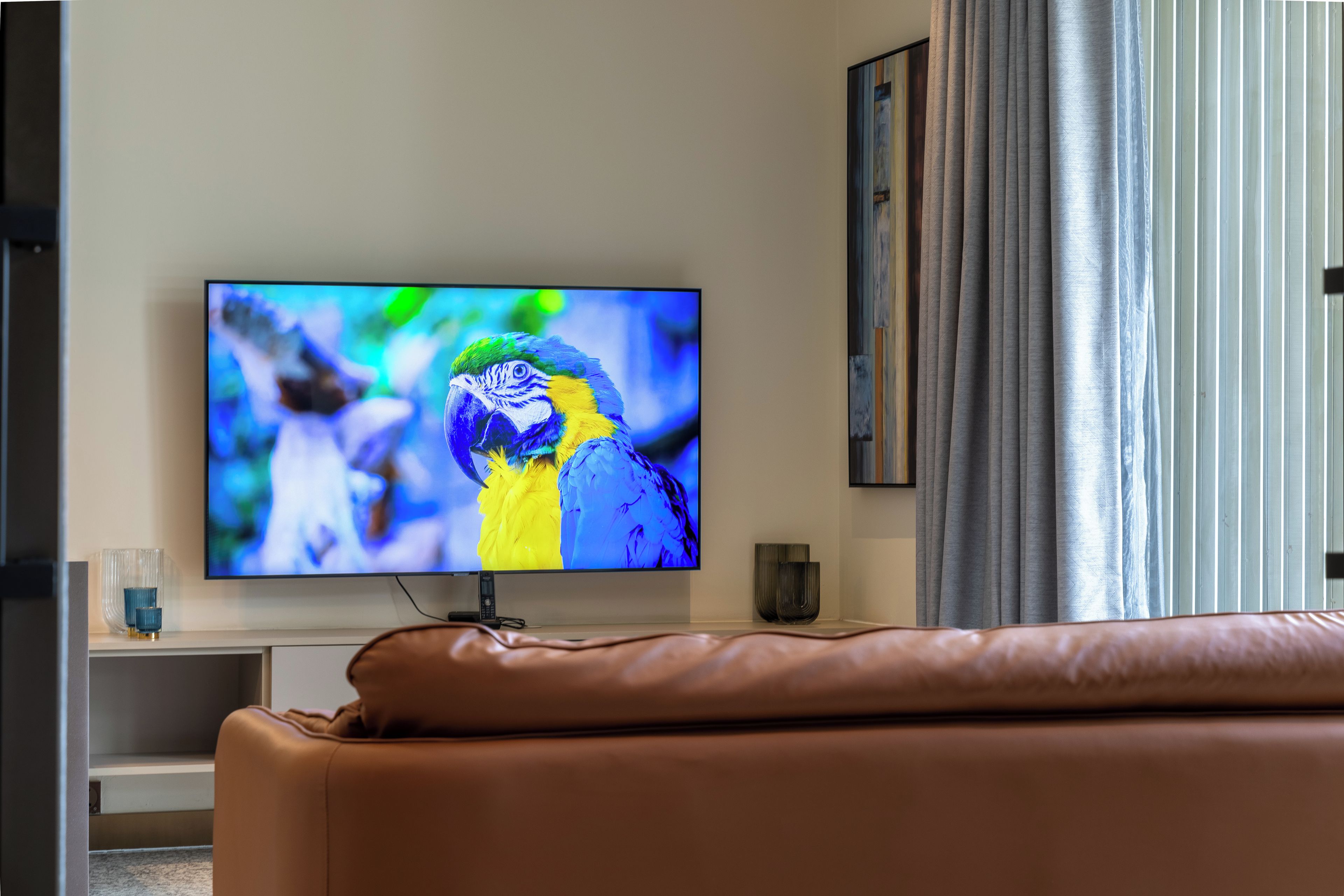 Televisão de tela plana fixada na parede, um sofá de couro marrom em primeiro plano e uma janela com cortinas à direita.