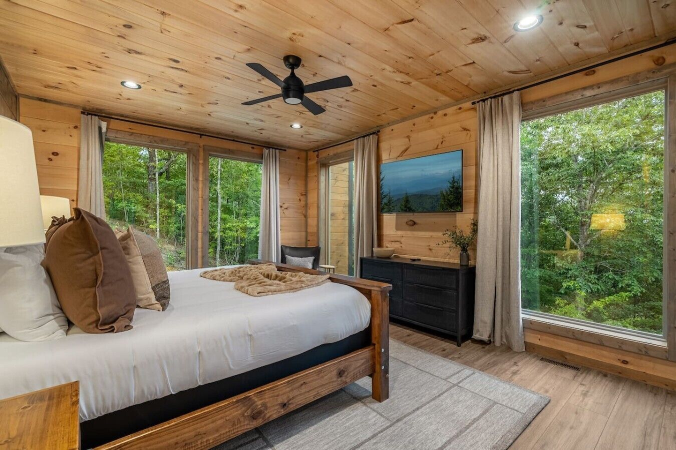 Un dormitorio con paneles de madera, una cama, ventilador de techo, televisor y ventanas amplias que ofrecen vistas al exterior.