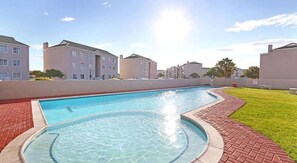 A swimming pool with a brick border, surrounded by a grassy area and residential buildings. - Private Room Near the Beach (Cape Town)
