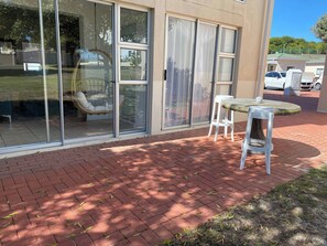 A patio area with a glass door, a wicker chair, and a small table. - Private Room Near the Beach (Cape Town)