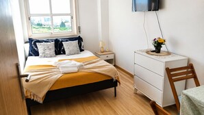 A bedroom with a bed, bedside table, dresser, chair, and a TV on the wall. - Room-singh Leite Holiday Home (Lisbon)