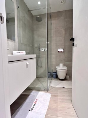 A modern bathroom with a glass shower enclosure, a white sink with a towel labeled "SuperHost," and a toilet with a trash bin beside it. - Le Bohémien Signature Apartment in La Marsa (La Marsa)