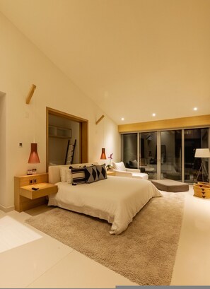 A modern bedroom with a large bed, bedside tables, a nightstand, a lamp, a window, and a sliding glass door leading to a balcony. - Casa Brisa 7BD - Kupuri Punta Mita (Punta de Mita)