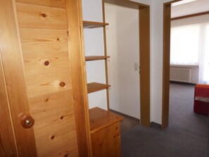 Ein Holzkleiderschrank mit mehreren Einlegeböden und einem kleinen Schrank, ein Durchgang zu einem anderen Raum sowie ein Heizkörper im Hintergrund.