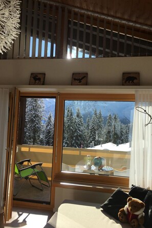 Habitació amb una finestra gran que dona a un paisatge de muntanya nevada, un balcó de fusta i una cadira plegable verda.