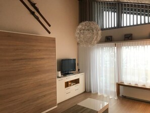 Un saló modern amb un armari de fusta ampli, un televisor de pantalla plana sobre un moble blanc, una làmpada penjant i cortines lleugeres.